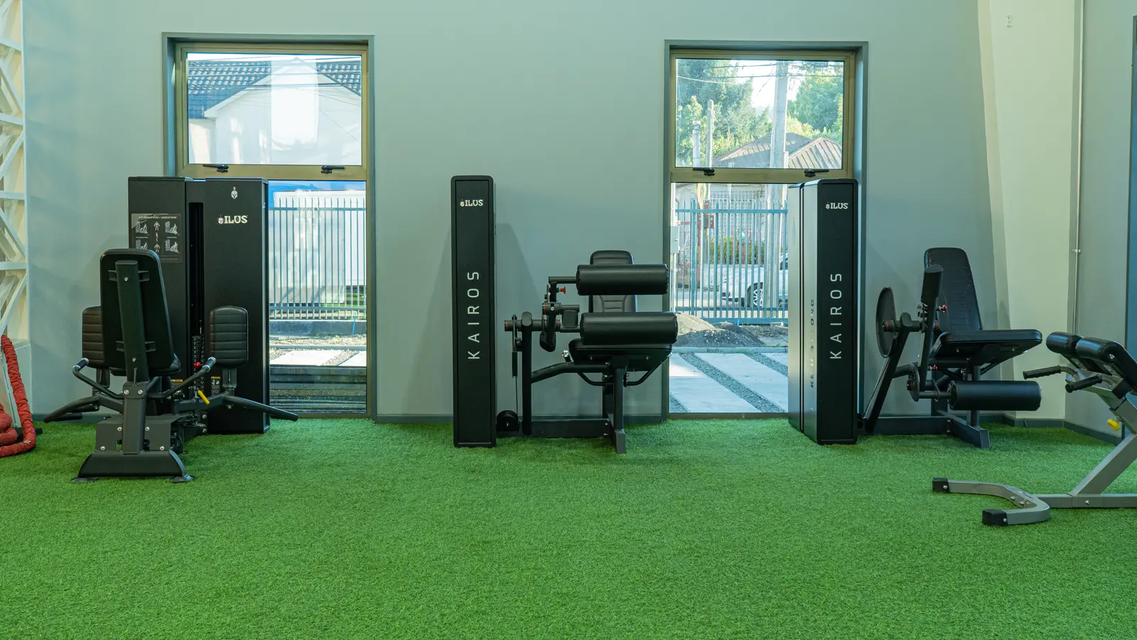 Interior de un gimnasio con suelo de césped artificial verde, máquinas de ejercicio contra una pared gris y ventanas. Espacio de entrenamiento luminoso, limpio y acogedor.