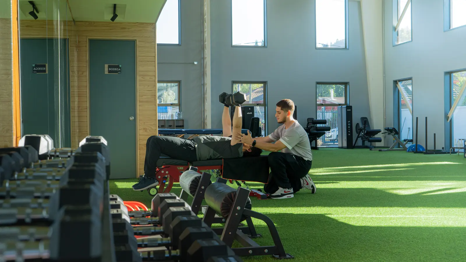 Un entrenador personal ayuda a un hombre a levantar mancuernas en un banco en un gimnasio luminoso con grandes ventanas y suelo verde.