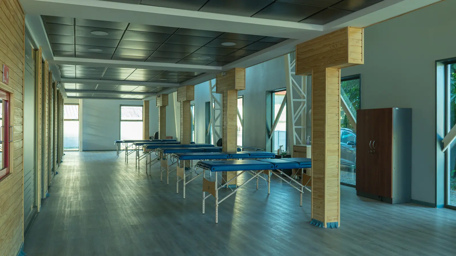 Una sala espaciosa con una fila de camillas de masaje azules vacías, pilares de madera y grandes ventanas que permiten la entrada de luz natural.