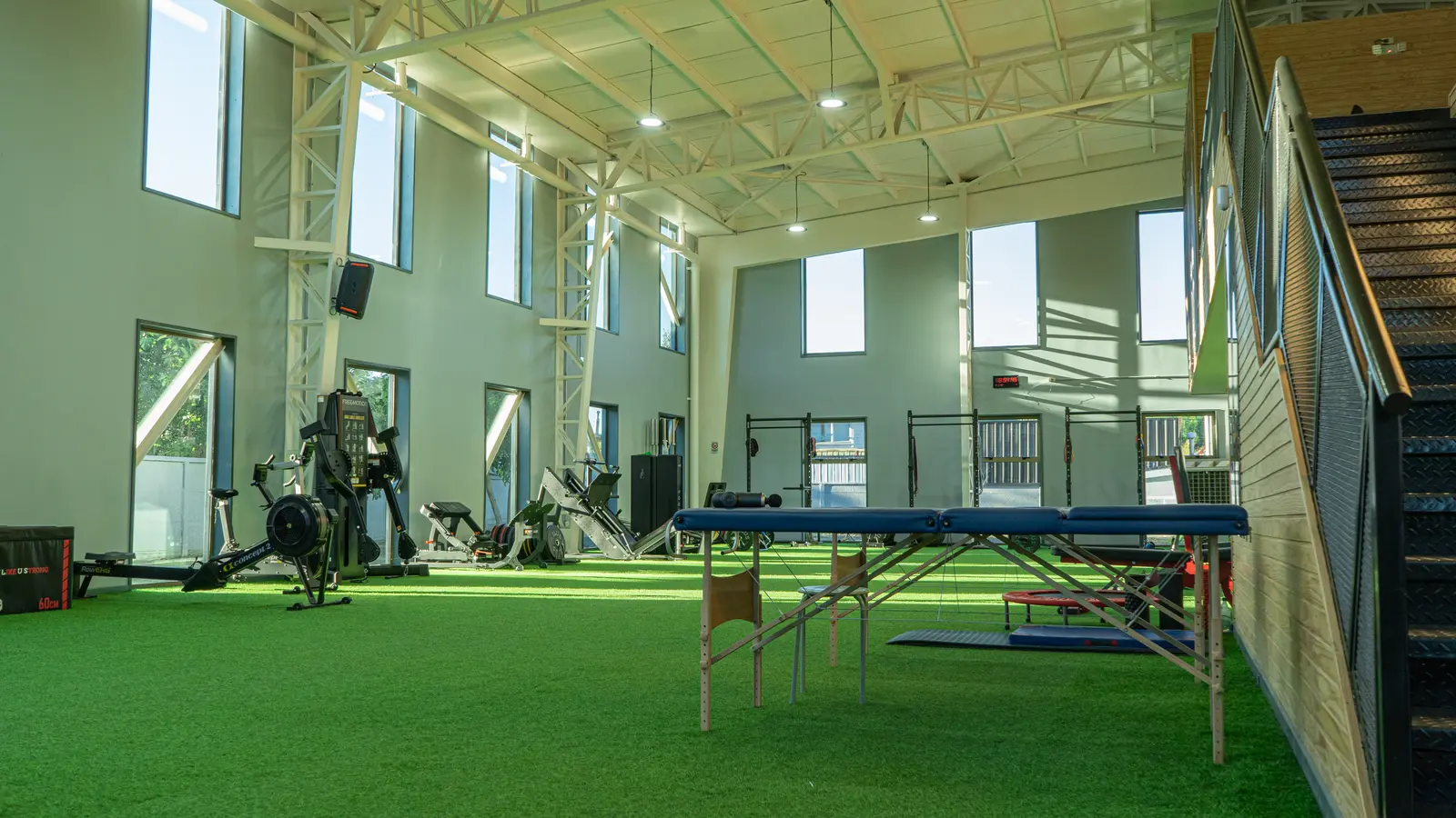 Gimnasio espacioso con techos altos, grandes ventanas y suelo de césped artificial. El equipamiento incluye máquinas de remo y pesas. Ambiente luminoso y aireado.