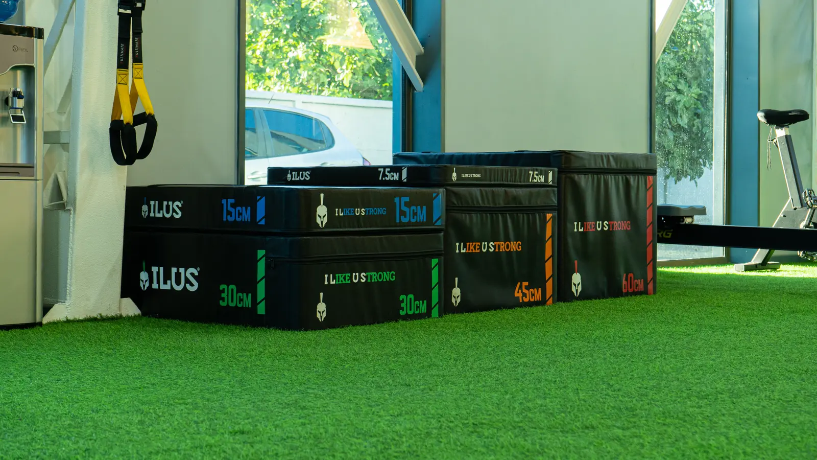 El gimnasio, con suelo de césped artificial, cuenta con cajas pliométricas apiladas etiquetadas con diferentes alturas. Cerca hay correas de suspensión amarillas y una bicicleta estática.