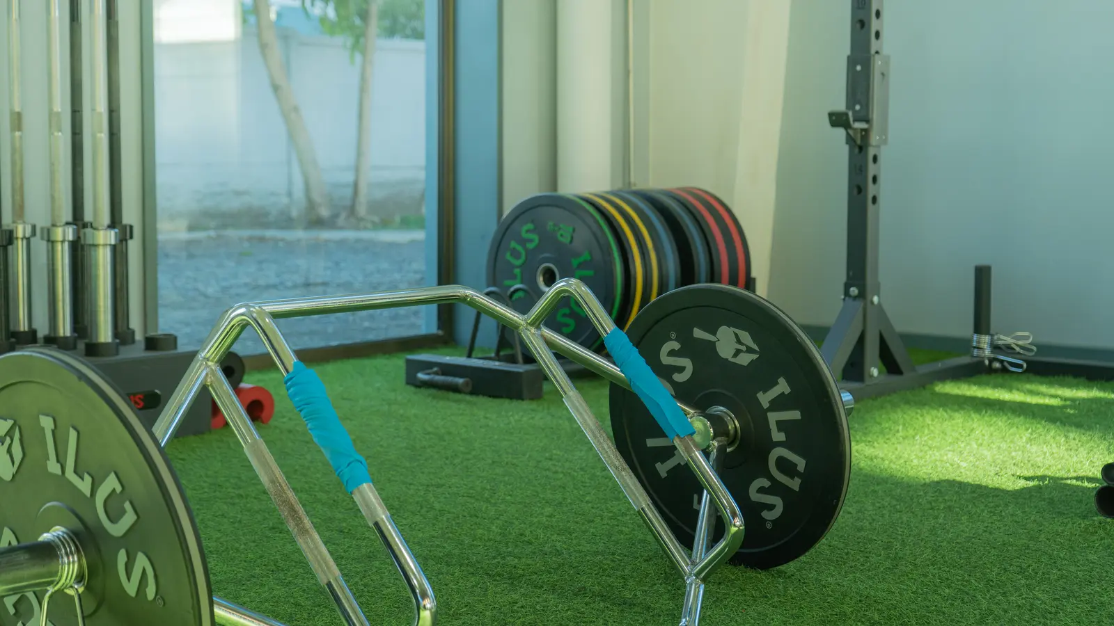 Una sala de gimnasio con equipamiento para levantamiento de pesas sobre un suelo de césped artificial verde. En primer plano se ve una barra hexagonal cargada, con discos y barras apilados cerca.