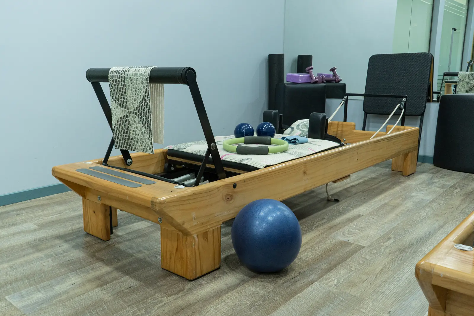 Una máquina Pilates Reformer en un estudio con una pelota azul de ejercicios y diversos accesorios de fitness cerca, sobre un suelo de madera.
